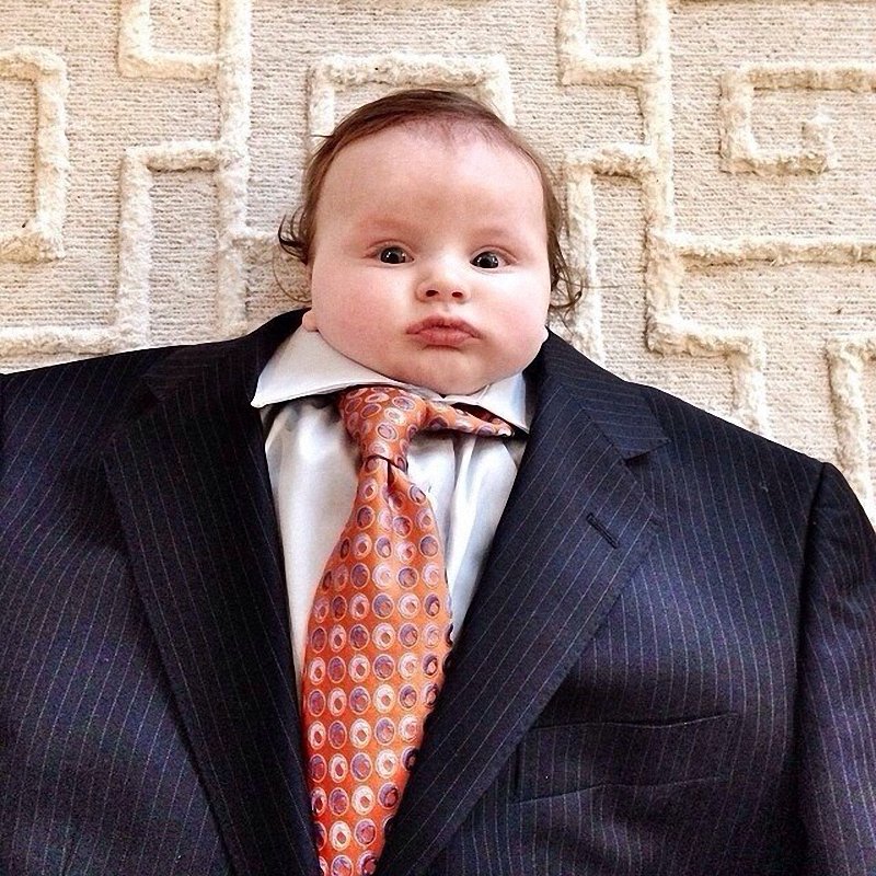 Маленький мальчик сидит одетый во взрослый костюм с галстуком.