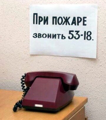 На столе стоит старый телефон без кнопок и без диска, а на стене весит объявление: При пожаре звонить 53-18.